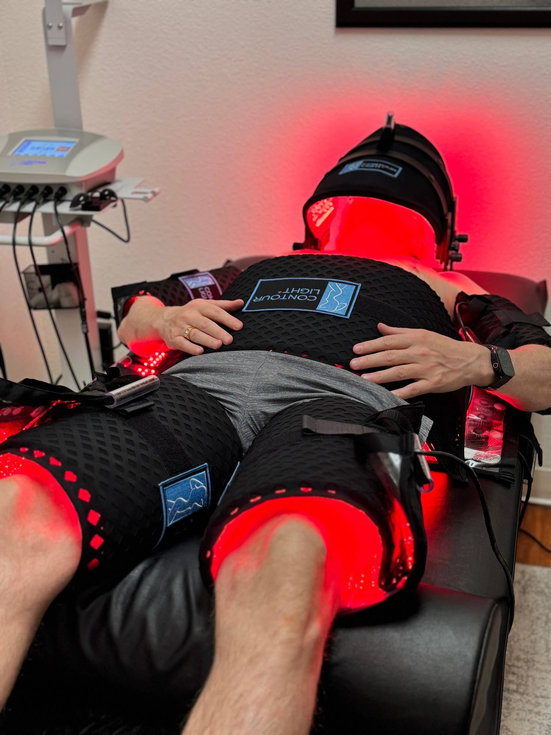 A client mid-session at Genesis Red Light in Cedar Park, TX: Contour Light pads glowing red around the waist, thighs and arms, face under the red LED panel