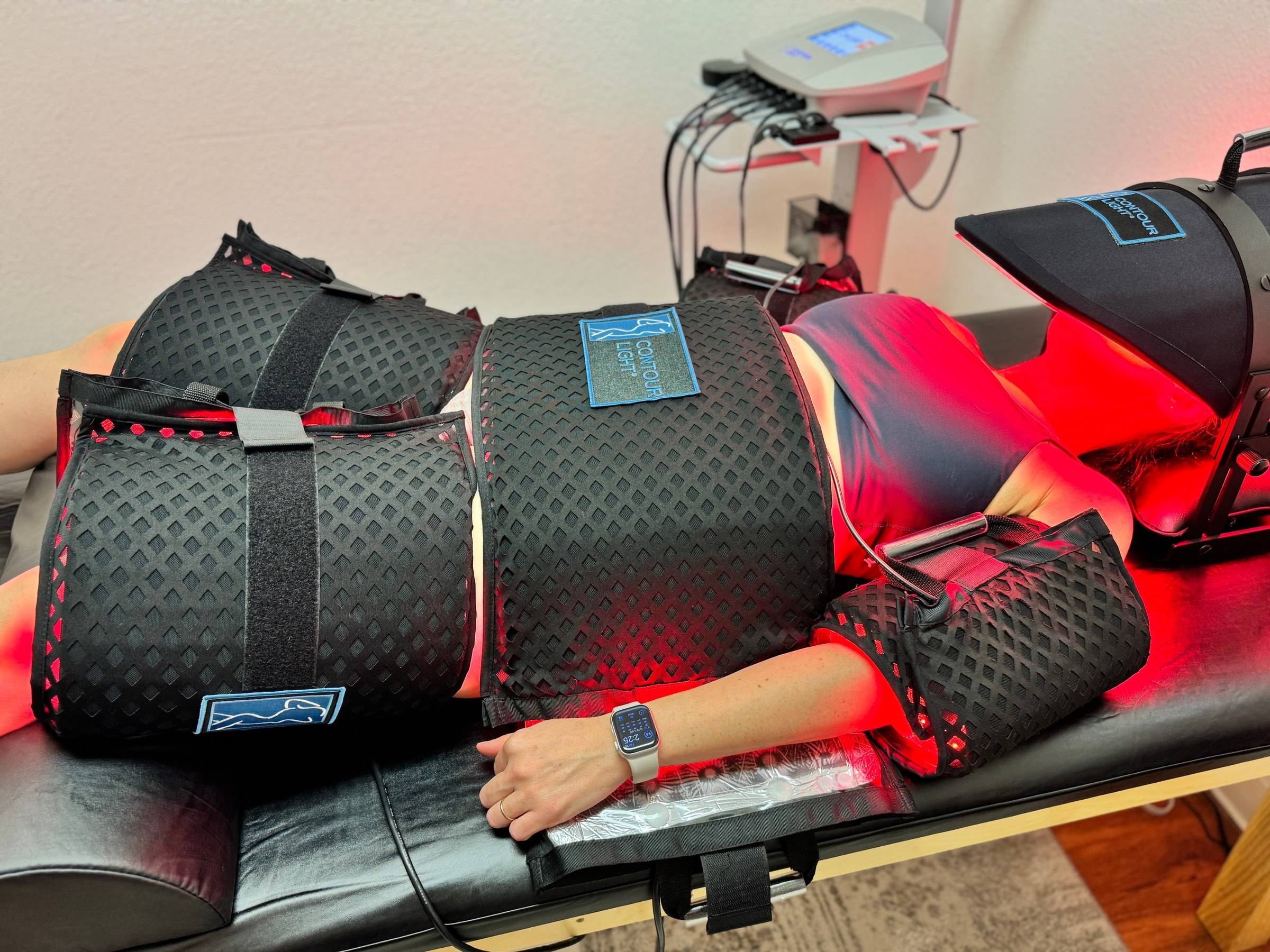 A client lying on the treatment bed, wrapped in black Contour Light pads around the waist, hips, thighs and arms, at Genesis Red Light in Cedar Park, TX