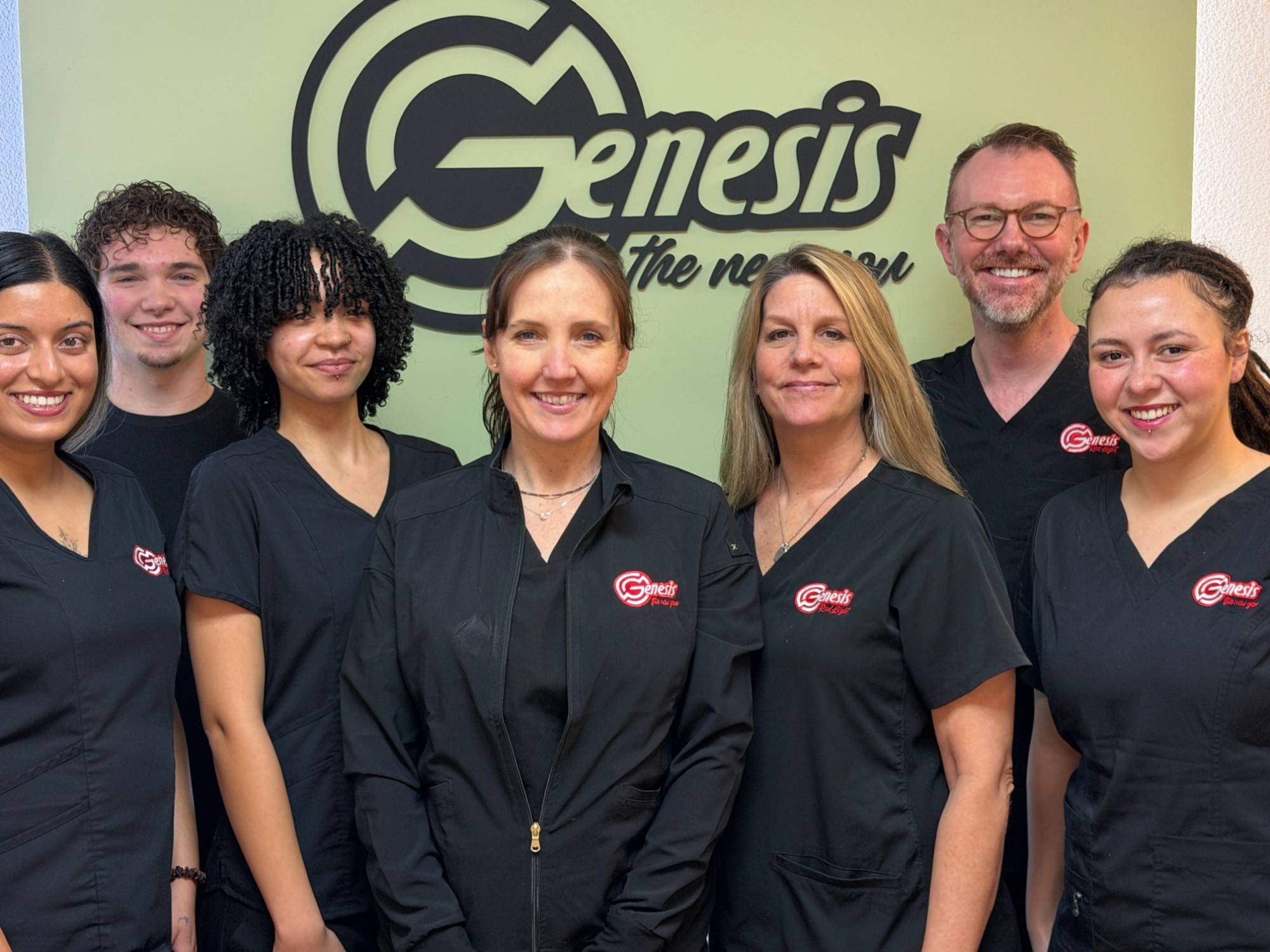 The Genesis Red Light team in black Genesis scrubs in front of the Genesis logo wall in the lobby in Cedar Park, TX. Left to right: Durga, Elijah, Alexa, Serenity, Tammy, John and Geraldine.